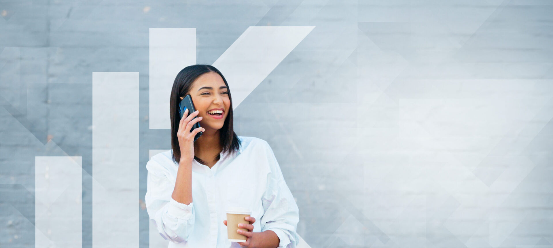 A woman wearing a white shirt smiles while talking on a smartphone and holding a coffee cup, standing against a gray wall with geometric white patterns.