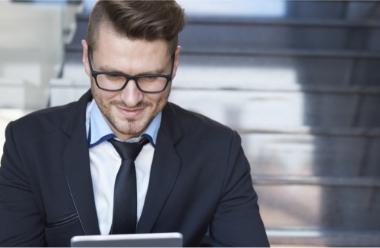 A man wearing glasses and a suit sits on stairs, smiling while looking at a tablet.