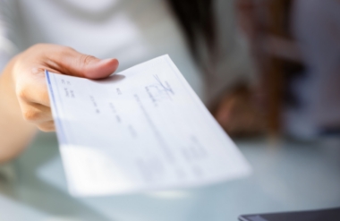 A person’s hand is holding out a paper check, offering it to someone else across a desk or table. The background is softly blurred.
