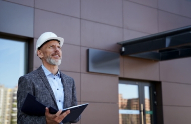 A smiling man wearing a white safety helmet and blazer holds a clipboard while standing outside a modern building. He looks upward, with part of the building and cityscape visible in the background.