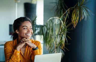 A woman in a mustard yellow shirt sits at a desk with a laptop, smiling and looking out the window. Green plants and soft natural light fill the modern office space around her.