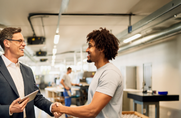 Two men smiling and shaking hands in a brightly lit industrial workspace, with one holding a tablet and others working in the background.