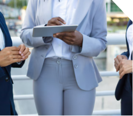 Three people in business attire stand outdoors, with one person holding a tablet and appearing to discuss something with the others. Their faces are not visible in the image.