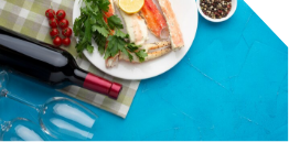 A plate of crab legs with lemon and parsley, cherry tomatoes, a bottle of red wine, two empty wine glasses, and a bowl of peppercorns on a blue table with a checkered cloth.