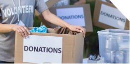 A person wearing a Volunteer shirt holds a box labeled Donations, with more donation boxes and supplies visible on a table in the background.